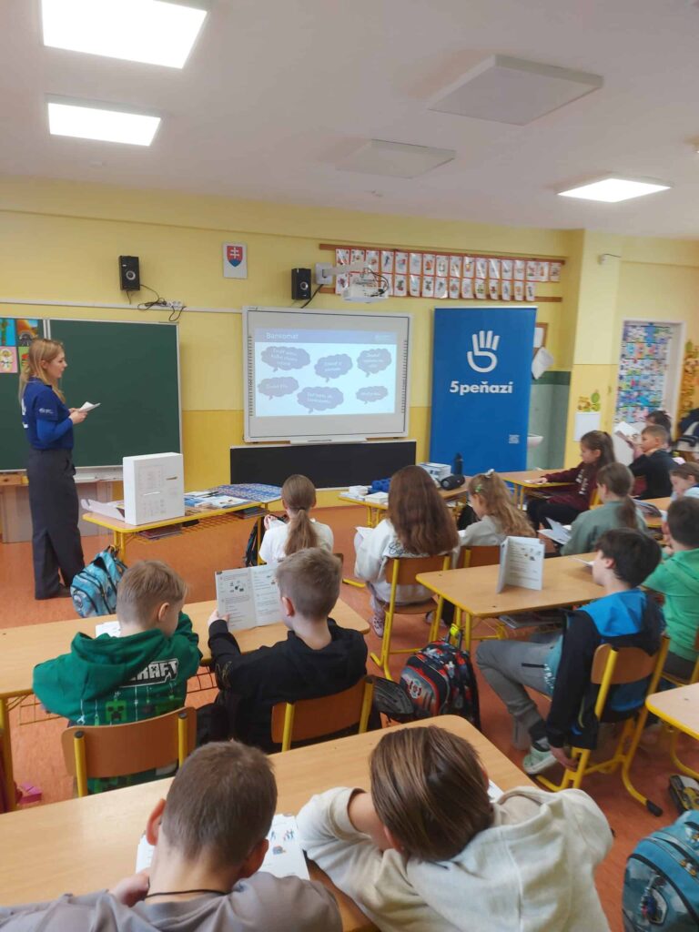 Učiteľ stojí vpredu triedy a prednáša skupine študentov sediacich v laviciach. Na obrazovke sa zobrazuje prezentácia s rečníckymi bublinami a vidno modrý transparent so symbolom bielej ruky.