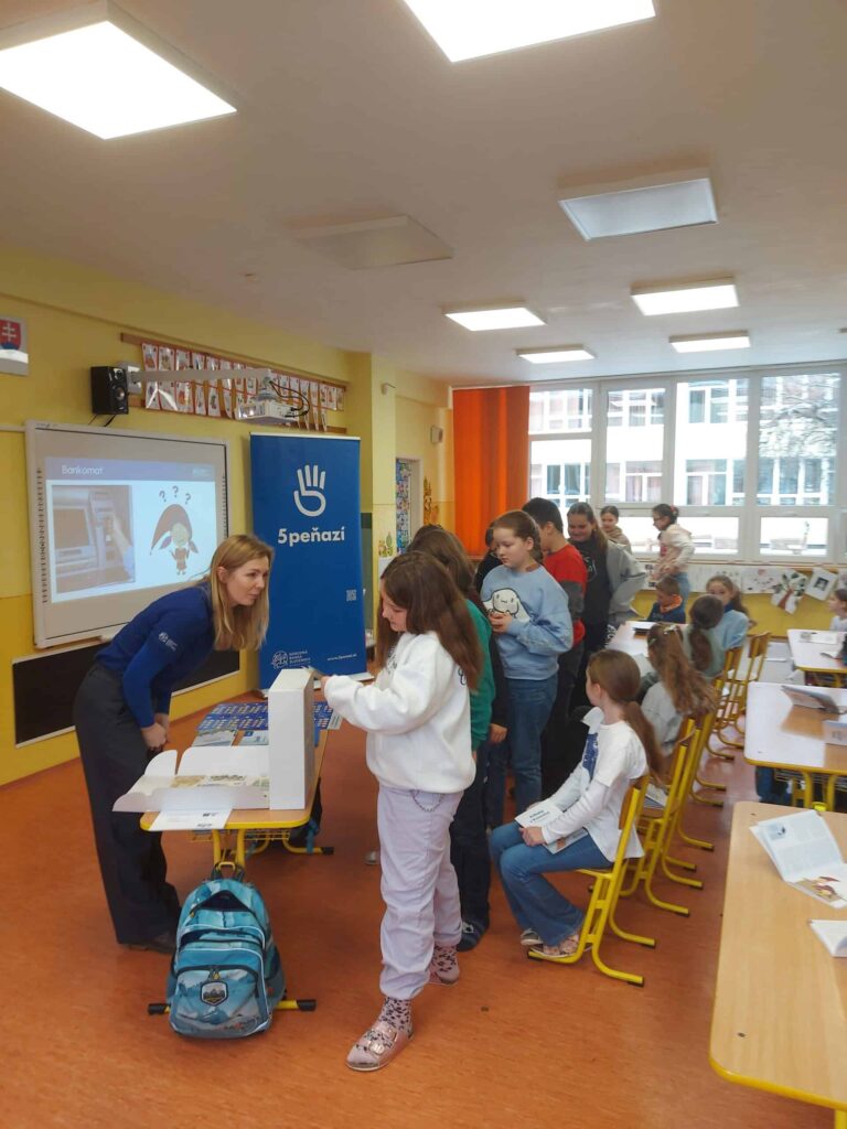Skupina detí sa postaví za stôl v jasne osvetlenej triede, kde s nimi komunikuje žena. Na obrazovke sa zobrazuje obrázok tučniaka a vedľa stojí modrý banner s textom a logom ruky.