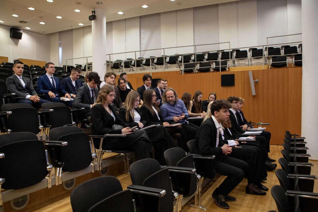 Skupina ľudí, väčšinou mladých ľudí vo formálnom oblečení, sedí v radoch v prednáškovej sále s poschodovými sedadlami a dreveným obložením. Niektorí sa pozerajú dopredu, iní používajú elektronické zariadenia.
