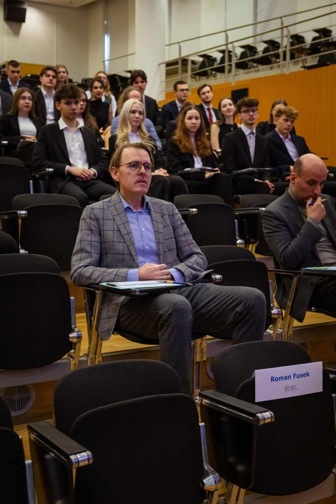 Muž v sivom kockovanom obleku sedí v prvom rade prednáškovej sály a ruky má zložené na stole. Za ním sedí niekoľko ľudí vo formálnom oblečení a pozorne ho počúvajú. Na tabuľke s menom je napísané Roman Fusek.
