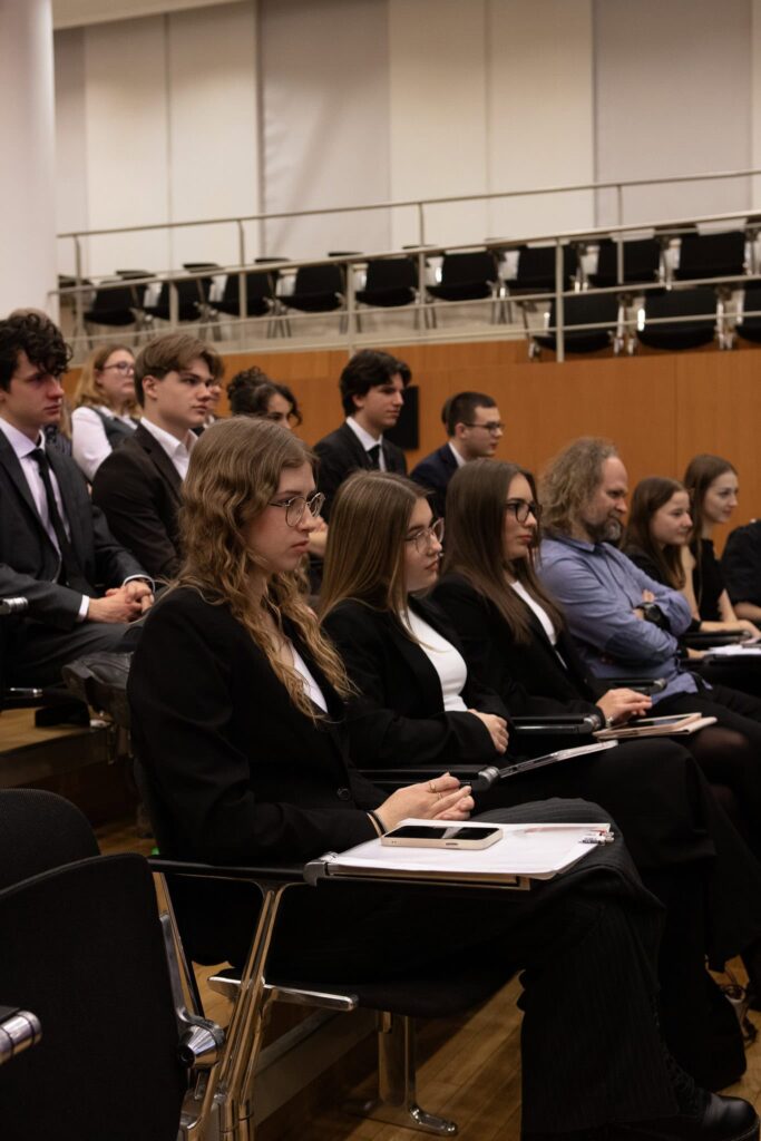 Skupina ľudí, väčšinou mladých ľudí vo formálnom oblečení, sedí pozorne v radoch stoličiek v prednáškovej sále alebo konferenčnej miestnosti a niektorí si robia poznámky.