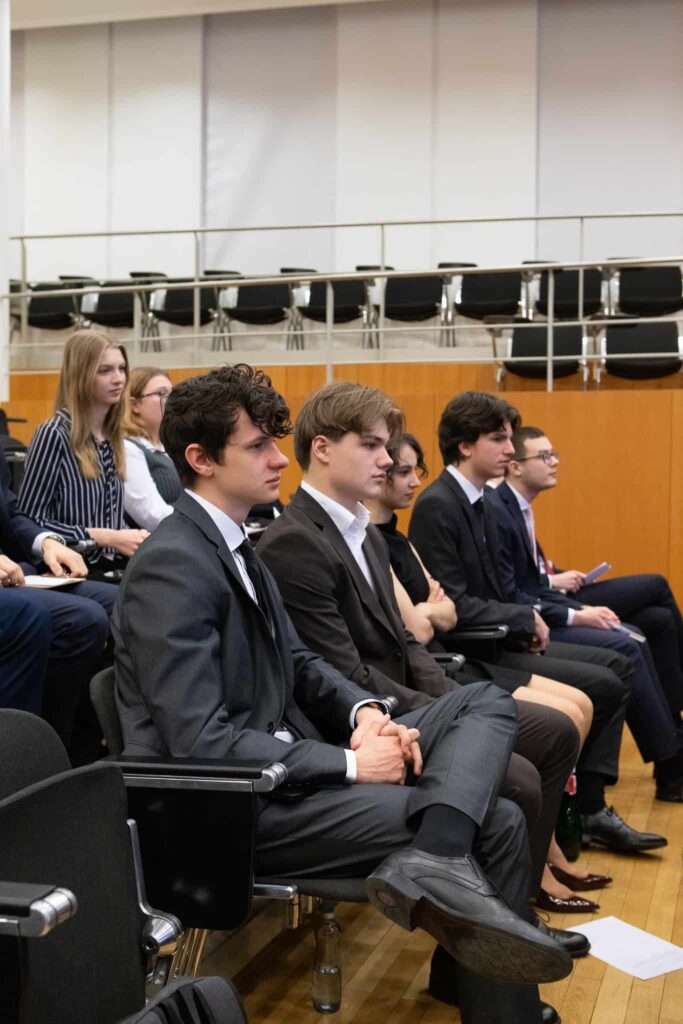 Skupina formálne oblečených mladých ľudí sedí pozorne v radoch čiernych stoličiek v prednáškovej sále, čo naznačuje, že sa zúčastňujú na prezentácii alebo konferencii.