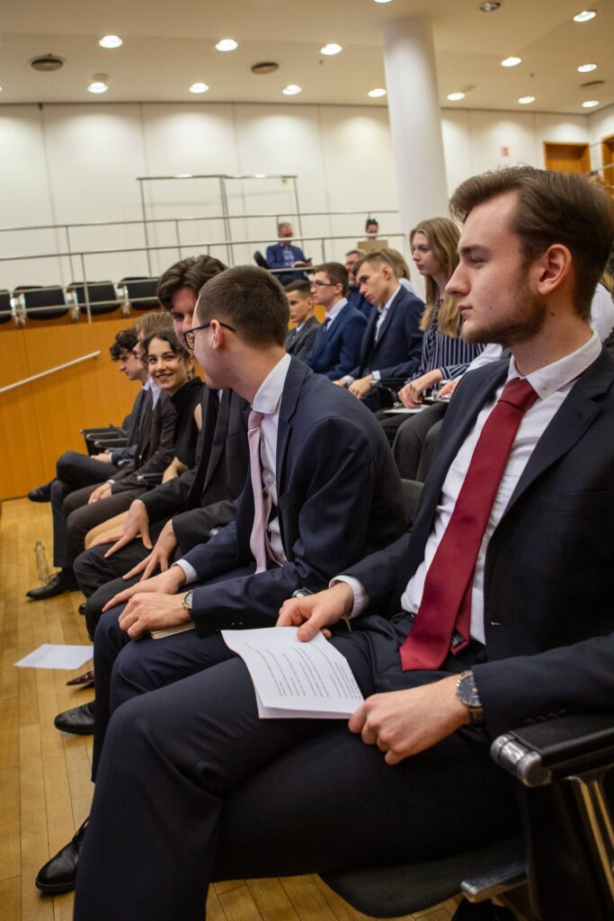 Skupina mladých ľudí vo formálnom oblečení sedí v radoch v modernej posluchárni, niektorí sa rozprávajú, iní počúvajú, v rukách majú papiere a okolo nich je niekoľko prázdnych miest.