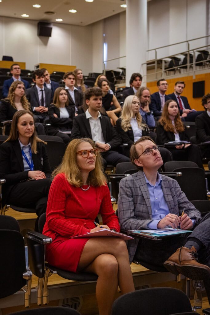 Skupina ľudí, väčšinou mladých ľudí vo formálnom oblečení, sedí v posluchárni, pozorne počúva a robí si poznámky počas konferencie alebo prednášky.