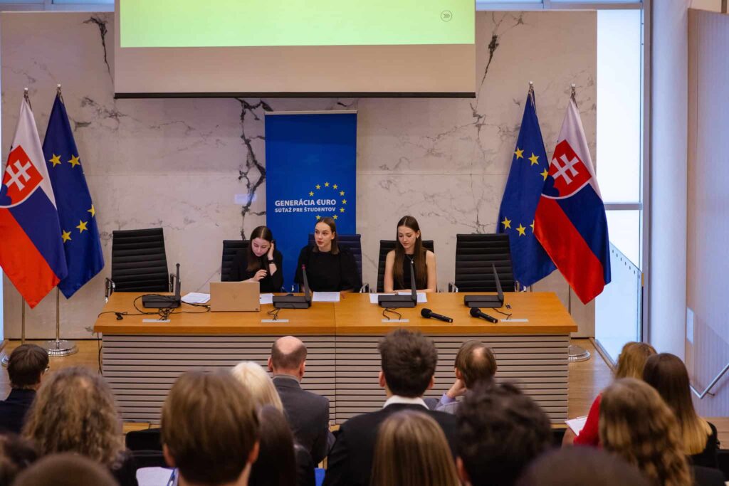 Tri ženy sedia za panelovým stolom pred publikom, za nimi sú zobrazené vlajky Slovenska a Európskej únie a nad nimi je prezentačné plátno. Miestnosť sa zdá byť pripravená na formálne podujatie alebo konferenciu.