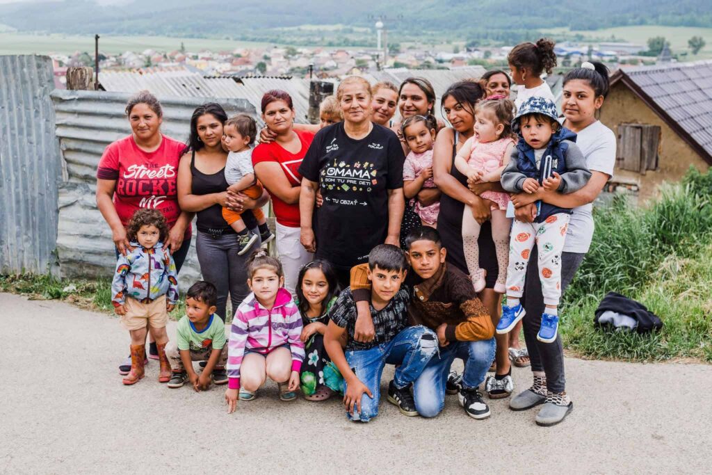 Skupina žien - Omám a detí spoločne pózuje vonku na vidieckej ceste, v pozadí vidno kovové oplotenie a domy a v diaľke dedinu.