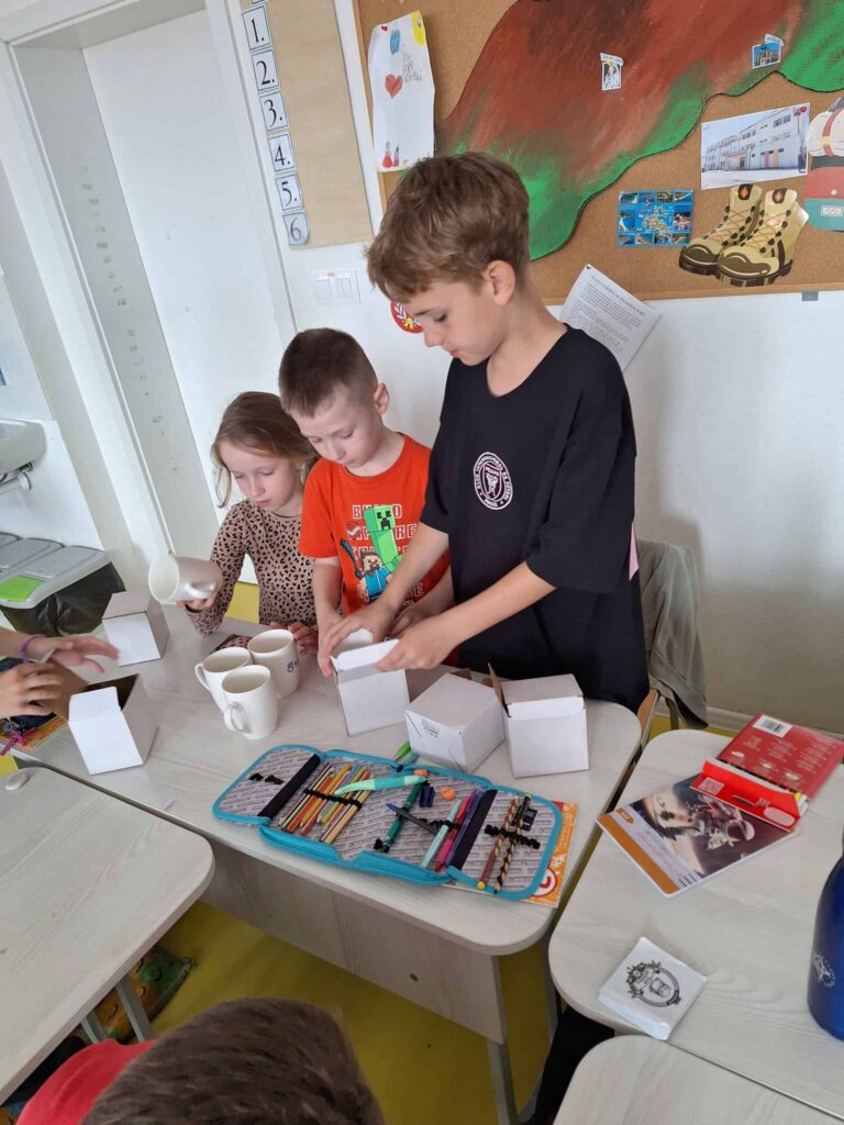 Tri deti stoja za stolom v triede a skladajú biele papierové škatule. Na stole sú rozhádzané výtvarné potreby, poháre a kresby. Na stene za nimi visí nástenka s farebnými výtvarnými prácami a papiermi.