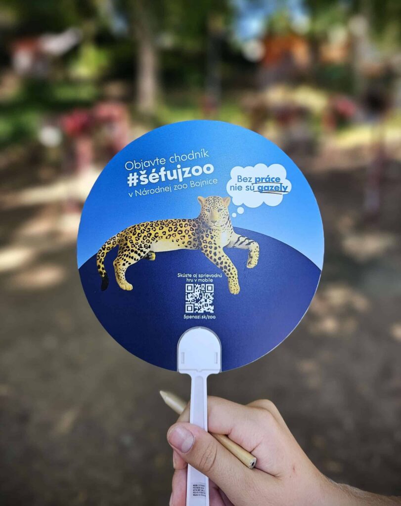 Ruka drží okrúhly vejár s ilustráciou leoparda, QR kódom a slovenským textom propagujúcim ZOO Bojnice. Pozadie je rozmazané s viditeľnou vonkajšou zeleňou.