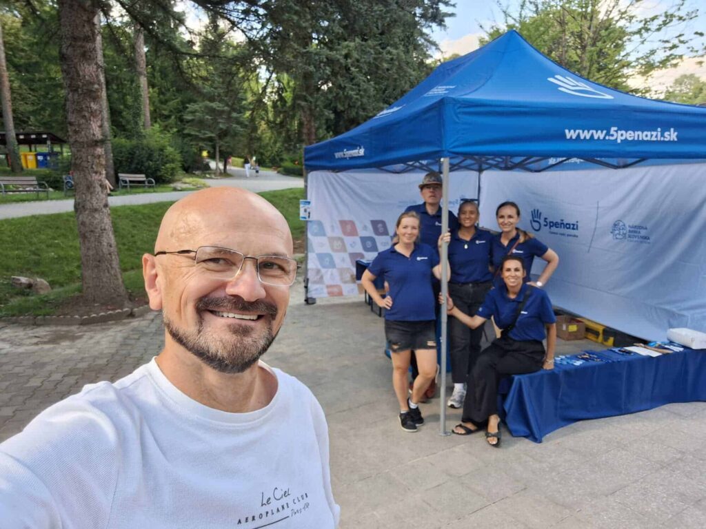 Usmievajúci sa muž si robí selfie pred modrým stanom, v ktorom stojí alebo sedí päť ľudí v rovnakých modrých tričkách, pózujú a usmievajú sa. Stan zobrazuje webovú stránku "5penazi.sk" a je postavený v parku so stromami a cestičkami.