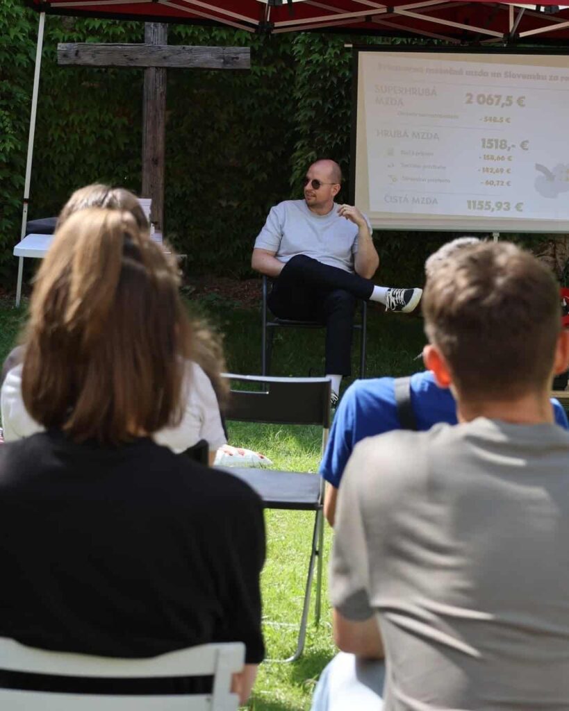 Muž sedí a hovorí k publiku pod stanom, za ním sa premieta prezentácia s údajmi o platoch v slovenčine. Publikum sedí na stoličkách a priestor obklopuje zeleň.