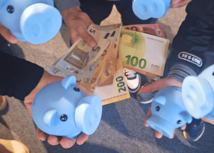 Niekoľko ľudí drží modré pokladničky a ukazuje eurobankovky vrátane bankoviek v hodnote 100 €, 200 €, 20 € a 5 €. Zameriavajú sa na sporenie a hospodárenie s peniazmi, pričom vidno ruky a ležérne oblečenie.