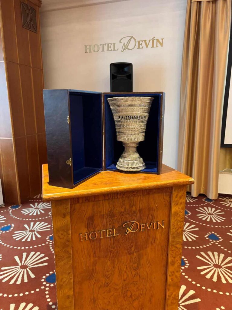 Na drevenom pódiu s vyrytým nápisom HOTEL DEVIN stojí veľká zdobená trofej vystavená v modrej zamatovej vitríne. V pozadí je na stene rovnaké logo HOTEL DEVIN a béžový záves.