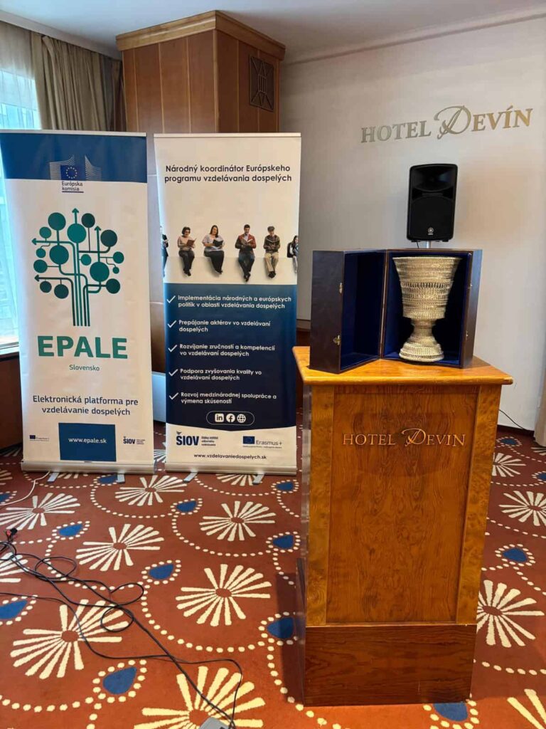 Pódium s trofejou a rečníkom stojí pred transparentmi EPALE Slovensko a Európskeho programu vzdelávania dospelých v hoteli Devín. Koberec je vzorovaný a vidno okno so závesmi.