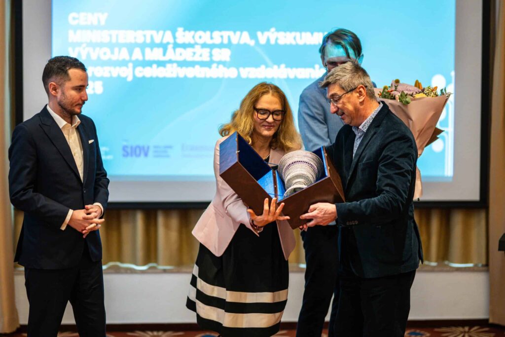 Štyria ľudia stoja spolu na slávnostnom odovzdávaní cien. Žena v čiernobielych šatách preberá od muža trofej a certifikát, zatiaľ čo sa na ňu pozerá ďalší muž a tretia osoba v pozadí drží kyticu kvetov.