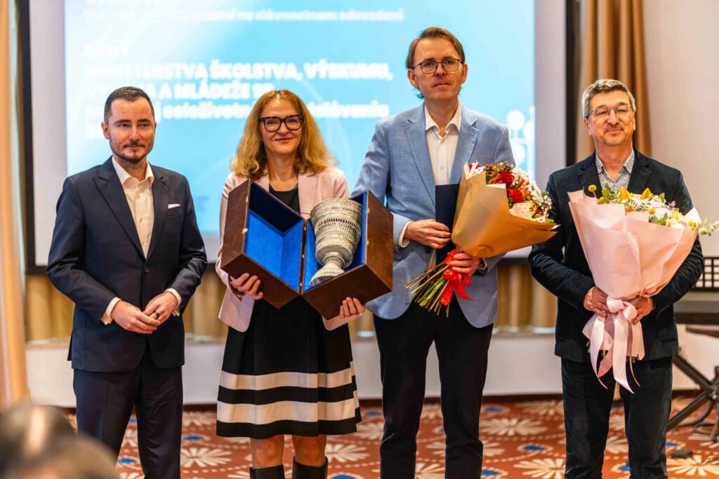 Štyria formálne oblečení ľudia stoja vedľa seba na slávnostnom odovzdávaní cien. Dvaja z nich držia kytice kvetov a jeden drží trofej v modrom puzdre. V pozadí je vidieť prezentačnú obrazovku.