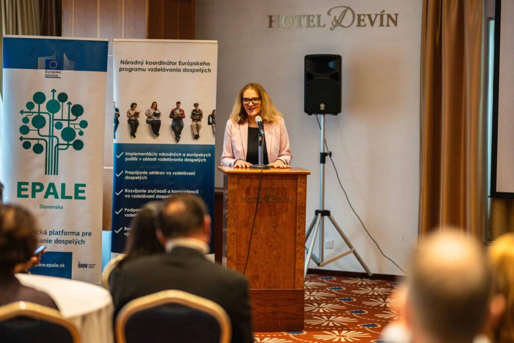 Žena stojí na pódiu a hovorí k poslucháčom v konferenčnej miestnosti, vedľa nej sú transparenty s logom EPALE Slovensko a informáciami. V pozadí je zobrazené logo HOTEL DEVÍN.