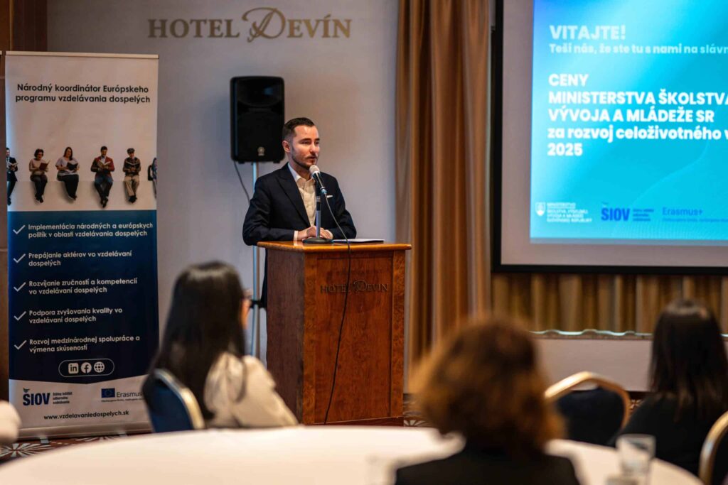 Muž v obleku stojí na pódiu a hovorí do mikrofónu v konferenčnej miestnosti. Za ním je transparent a premietacie plátno, na ktorom sa zobrazuje text v slovenčine. V popredí sú viditeľné okrúhle stoly a stoličky.
