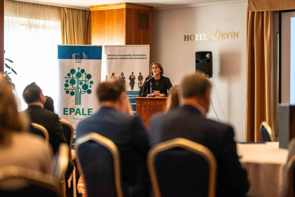 Žena reční na pódiu v konferenčnej miestnosti so sediacimi účastníkmi. Za ňou sú transparenty EPALE a Európskej únie. Prostredie je hotel Devín a vidno premietacie plátno.