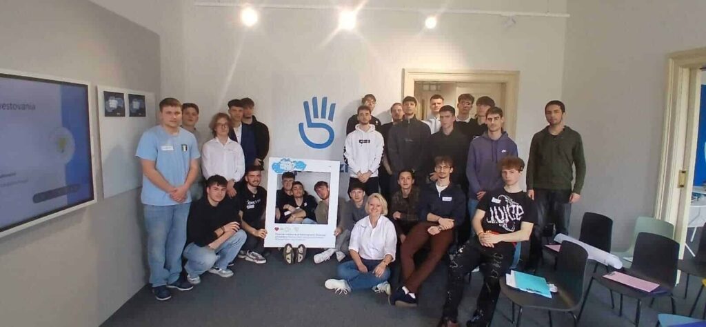 Skupina asi 23 mladých dospelých a jedna staršia žena pózujú v interiéri so symbolom veľkej ruky a obdĺžnikovou fotoprípravou; niektorí stoja, iní sedia alebo kľačia, všetci sú otočení tvárou k fotoaparátu.