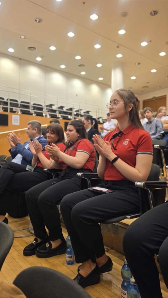 Skupina žiakov v červených tričkách sedí v sále a pozorne tlieska v NBS počas finále Peňažného štvorboja.