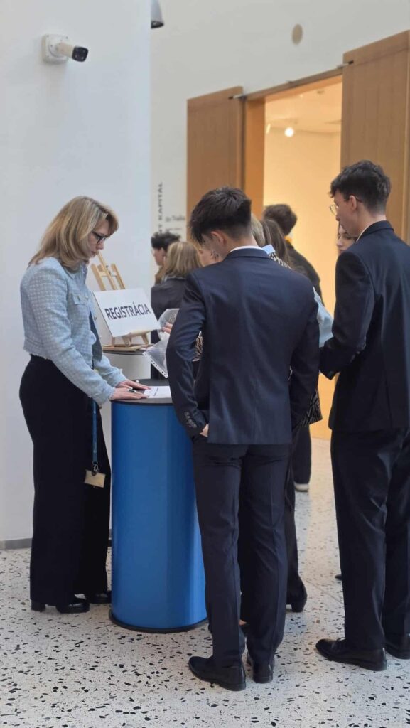 Žena stojí za registračným pultom a pomáha niekoľkým ľuďom vo formálnom oblečení pri registrácii na finále Peňažného štvorboja. Na stole je viditeľný nápis REGISTRÁCIA.