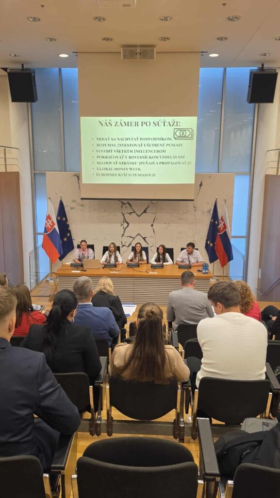 Skupina piatich žiakov sedí za stolom v konferenčnej miestnosti NBS a hovorí k publiku. Na oboch stranách sú vyvesené slovenské vlajky a za nimi sa premieta prezentácia so slovenským textom.