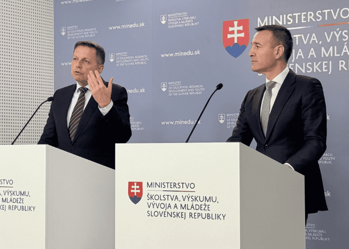 Dvaja muži v oblekoch stoja na pódiu s logom slovenského ministerstva školstva a reční na tlačovej konferencii. Jeden muž gestikuluje rukou, zatiaľ čo druhý sa pozerá dopredu. Na modrom pozadí sú zobrazené informácie slovenskej vlády.
