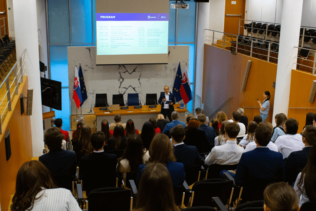 Rečník sa prihovára poslucháčom sediacim v prednáškovej sále s mramorovým pozadím, dvoma slovenskými vlajkami a prezentáciou premietanou na plátne nad ním. Účastníci sú otočení dopredu a po strane stojí žena, ktorá fotografuje.