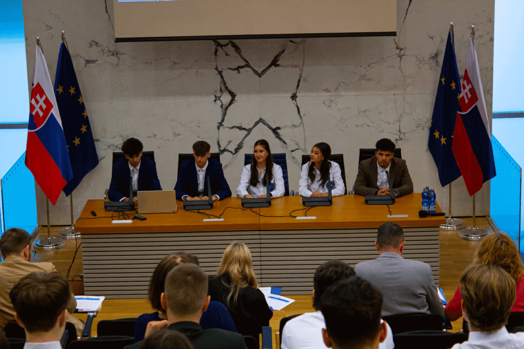 Päť mladých ľudí, tri ženy a dvaja muži, sedia za dlhým stolom s mikrofónmi pred publikom. Za nimi v slávnostnej konferenčnej miestnosti sú vyvesené slovenské vlajky a vlajky EÚ.