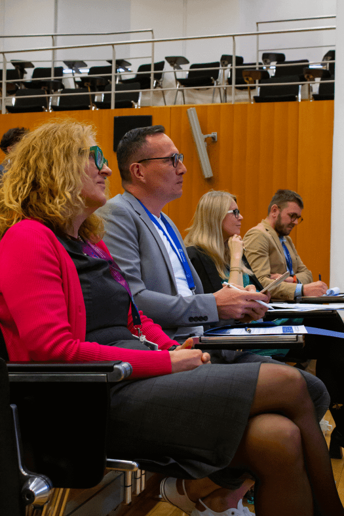 Štyria ľudia sedia v rade na konferencii alebo stretnutí, pozorne počúvajú a robia si poznámky. Majú na sebe pracovné oblečenie a konferenčné odznaky. Prostredie je moderná miestnosť s dreveným obložením a prázdnymi stoličkami v pozadí.