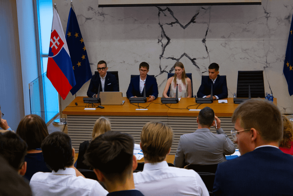 Štyria ľudia vo formálnom oblečení sedia za panelom s mikrofónmi pred publikom. Za nimi sú vlajky Slovenska a Európskej únie a mramorová stena, nad ktorou je premietacie plátno.