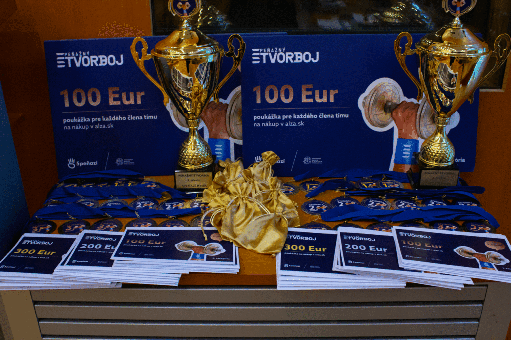 Na stole sú vystavené dve zlaté trofeje, medaily s modrými stuhami a niekoľko certifikátov na peňažné ceny (100, 200 a 300 eur), v pozadí je slovenský text a značka podujatia.