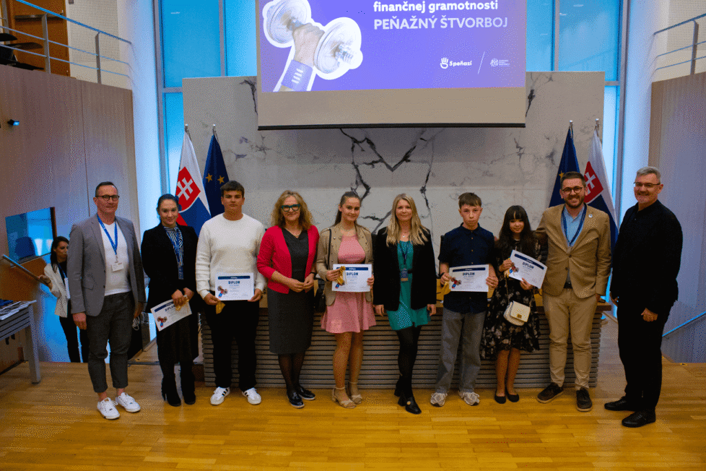 Skupina ľudí, vrátane študentov a dospelých, stojí vo vnútri a drží certifikáty. Za nimi sú dve slovenské vlajky a veľká prezentačná obrazovka so slovenským textom týkajúcim sa finančnej gramotnosti.