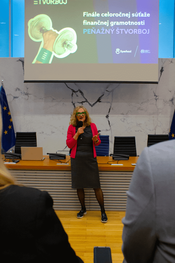 Žena v sivých šatách a ružovom saku hovorí do mikrofónu v konferenčnej miestnosti. Za ňou sa na obrazovke zobrazuje slovenský text a vlajky Európskej únie.