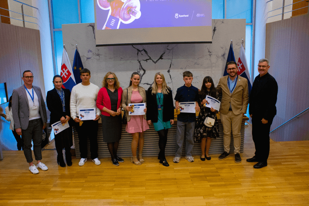 Skupina desiatich ľudí stojí v interiéri pred pódiom a v rukách drží certifikáty, po oboch stranách sú slovenské vlajky. Sú oblečení v elegantnom neformálnom oblečení a usmievajú sa do kamery.