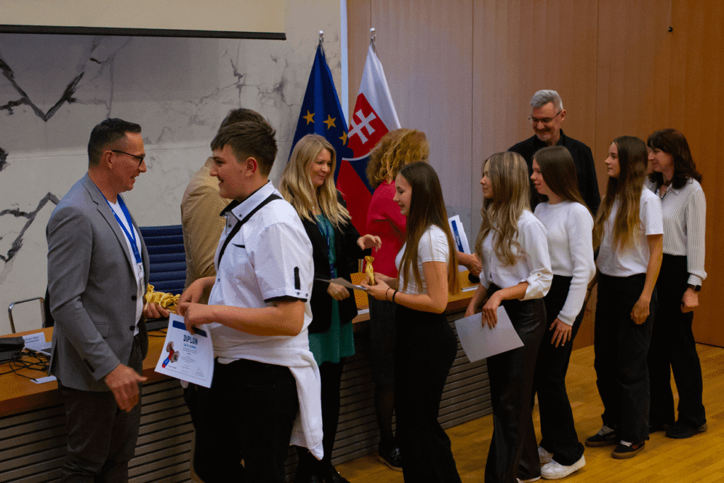 Skupina mladých ľudí, z ktorých niektorí držia certifikáty, stojí v rade, aby pozdravila alebo zablahoželala dospelým v slávnostnej miestnosti so slovenskými vlajkami a vlajkami EÚ za nimi.