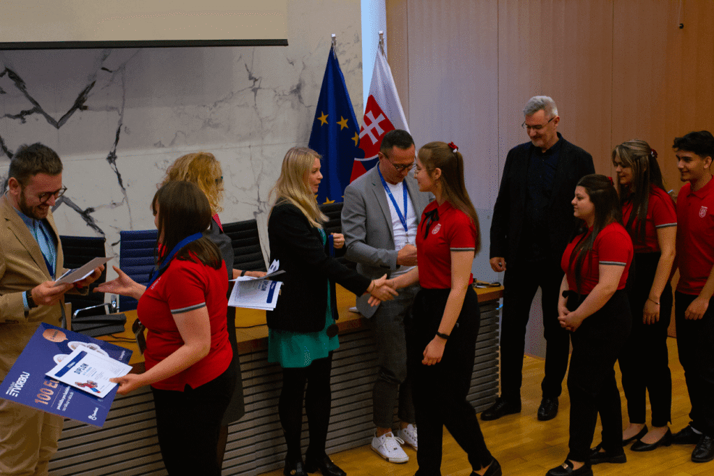 Skupina mladých ľudí v rovnakých červených tričkách dostáva certifikáty a podáva si ruky s úradníkmi v slávnostnej miestnosti s mramorovými stenami, vlajkami a drevenou podlahou. Zdá sa, že ide o slávnostné odovzdávanie cien alebo promócie.
