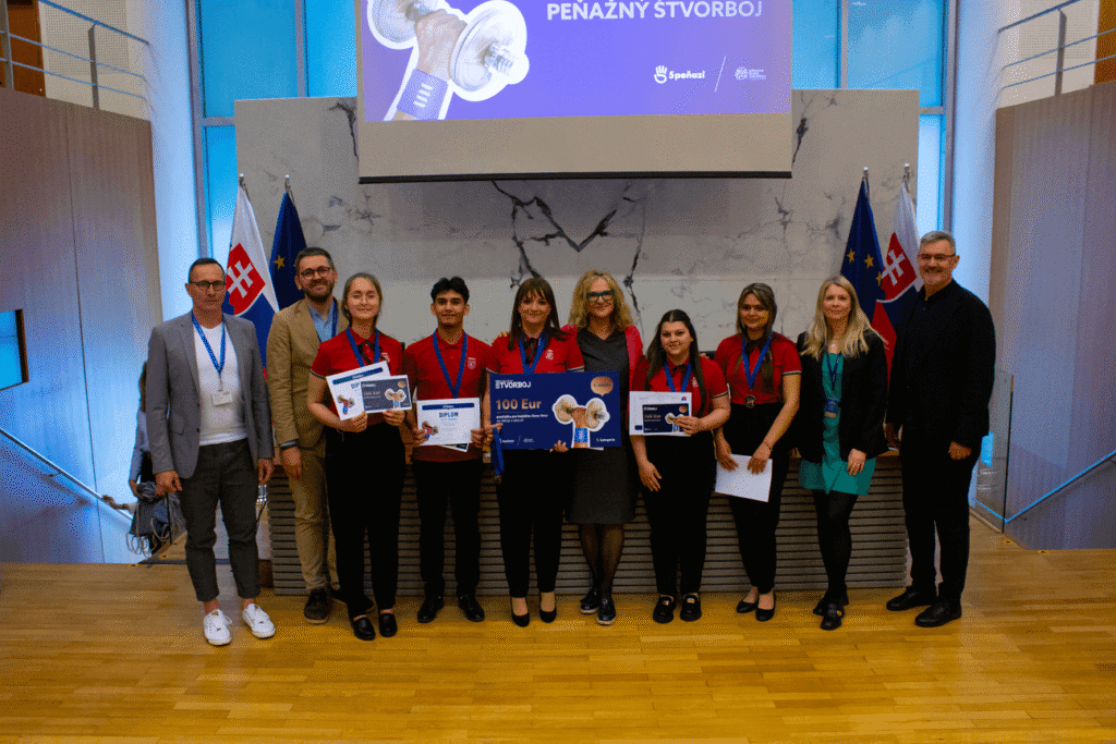 Skupina ľudí, niektorí s diplomami a cenami, stojí spoločne v interiéri na drevenej podlahe so slovenskými vlajkami a nápisom Penazny Stvorboj v pozadí. Skupina sa usmieva a pózuje na fotografiu.