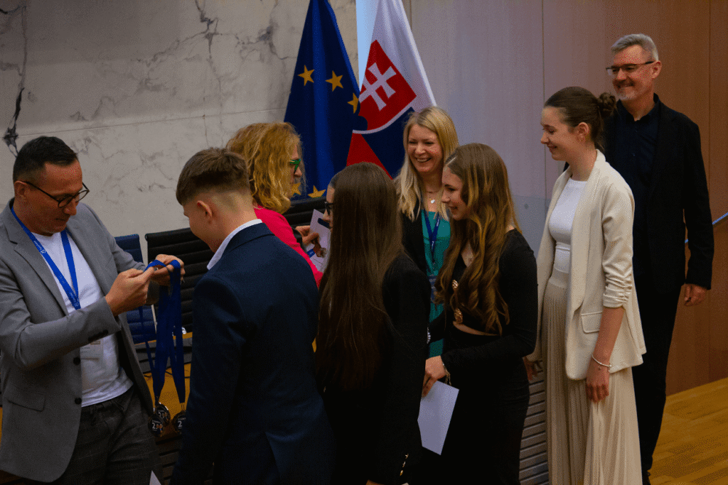 Skupina ľudí, z ktorých niektorí držia medaily a dokumenty, stojí v rade v interiéri pri vlajkách EÚ a Slovenska. Dvaja muži a štyri ženy sa usmievajú, sú oblečení vo formálnom oblečení, v prostredí slávnostného podujatia.
