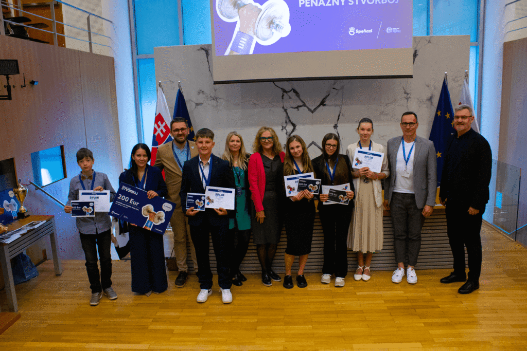 Skupina mladých ľudí a dospelých, z ktorých niektorí držia certifikáty a ceny, stojí spolu v slávnostnej miestnosti s drevenými podlahami, vlajkami a mramorovou stenou a pózuje na skupinovú fotografiu pri udeľovaní ocenení.