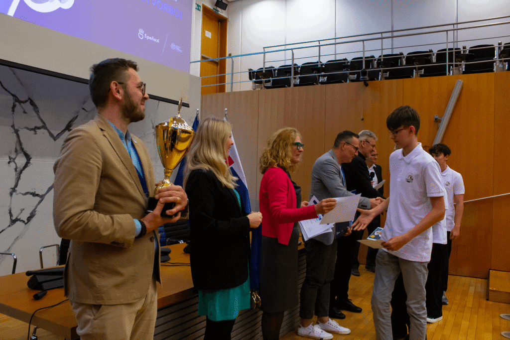 Skupina ľudí stojí v rade v modernej posluchárni. Jedna osoba drží trofej, zatiaľ čo ostatní odovzdávajú certifikáty študentom v bielych tričkách. V pozadí je vidieť stoličky a veľkú obrazovku.