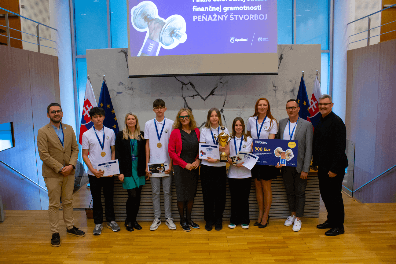 Skupina ľudí, medzi ktorými sú aj študenti s medailami, stojí v rade s oceneniami a veľkým šekom na 300 eur a pózuje na fotografiu v interiéri so slovenskými vlajkami a transparentom s cenami za nimi.