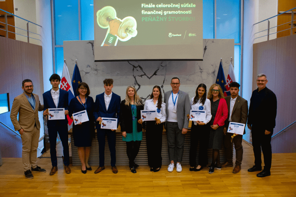 Skupina desiatich ľudí stojí v interiéri, drží certifikáty a medaily pred slovenskými vlajkami a vlajkami EÚ. Za nimi sa na obrazovke zobrazuje slovenský text o finále súťaže finančnej gramotnosti.