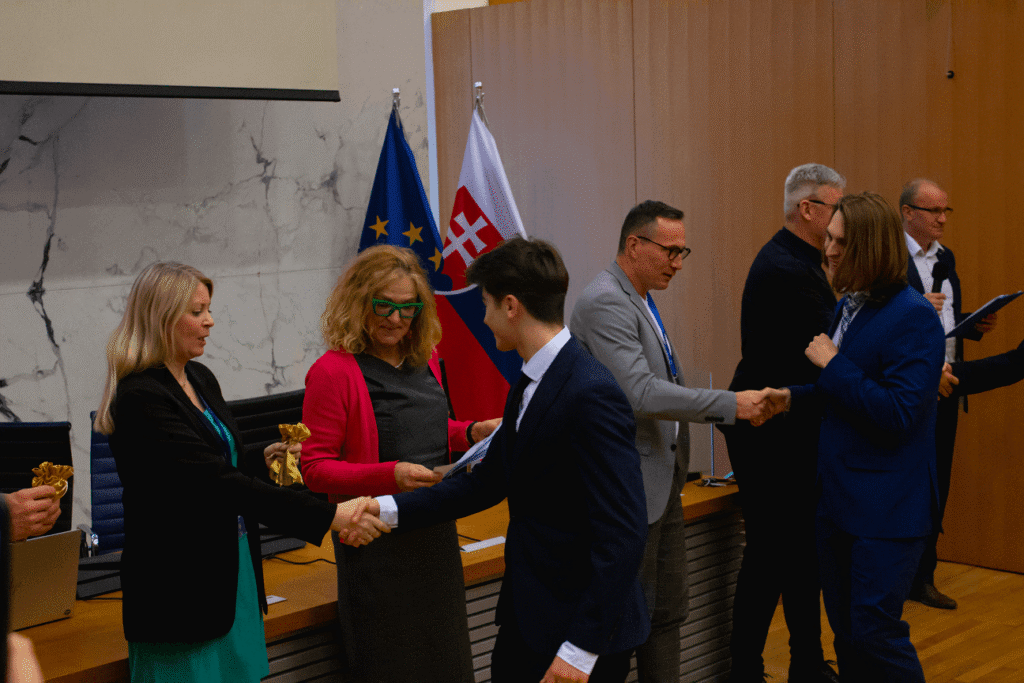 Niekoľko ľudí vo formálnom oblečení si v konferenčnej miestnosti podáva ruky a vymieňa si ocenenia. V pozadí vidno vlajky Európskej únie a Slovenska. Prostredie pôsobí oficiálne a slávnostne.