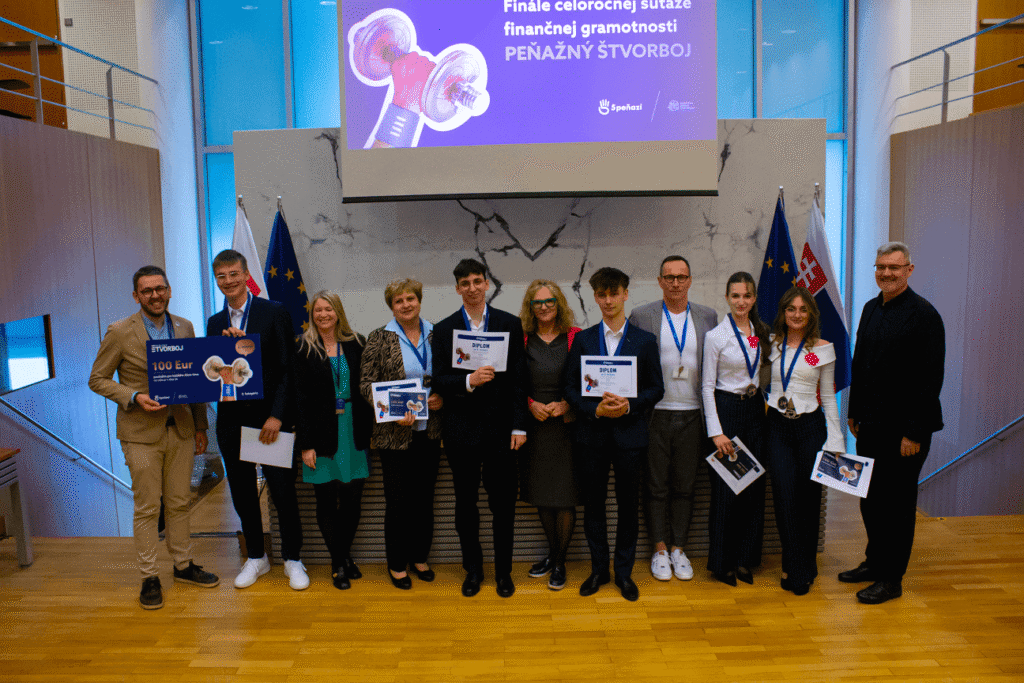 Skupina ľudí, z ktorých niektorí držia certifikáty a medaily, stojí v rade na drevenej podlahe pred vlajkami a veľkou obrazovkou vo formálnom prostredí, pravdepodobne na oslave nejakého ocenenia alebo súťaže.