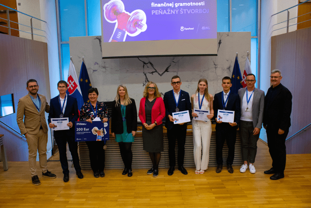 Skupina desiatich ľudí pózuje v interiéri; niektorí držia certifikáty a nadrozmerný šek. Za nimi sú slovenské vlajky a vlajky EÚ, nad hlavou sa na fialovej obrazovke zobrazuje text v slovenčine.