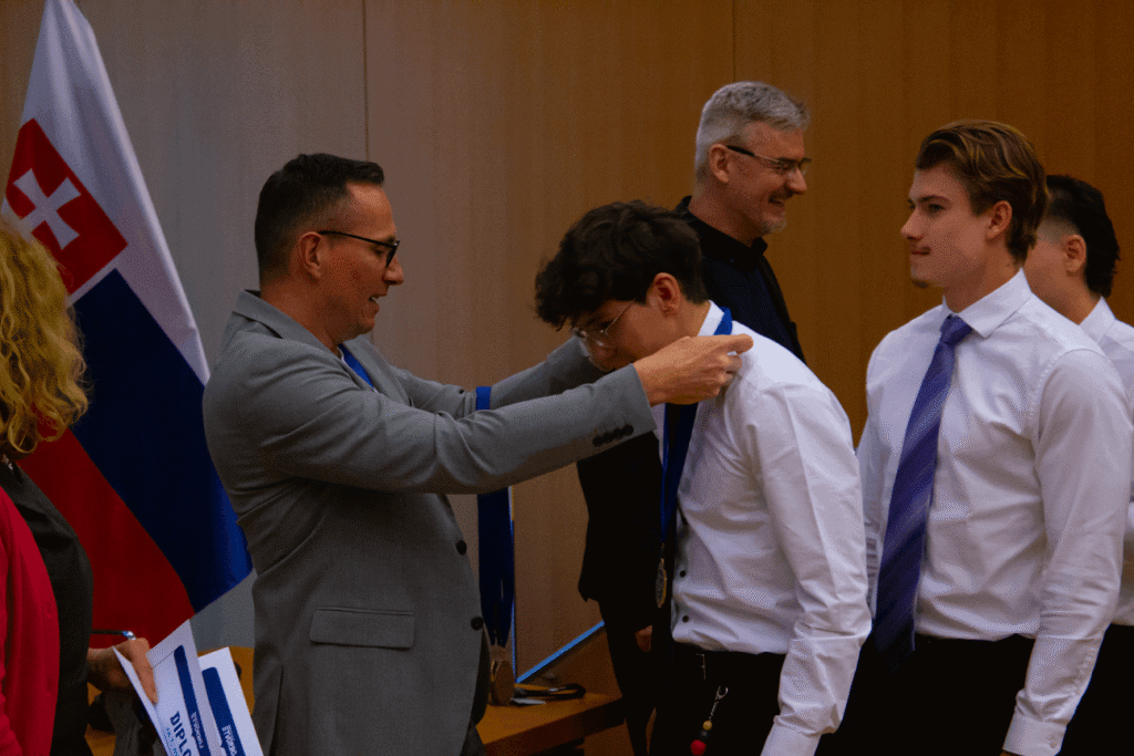 Muž v sivom obleku dáva na slávnostnom odovzdávaní cien medailu na krk mladému mužovi v bielej košeli. Neďaleko stoja ďalší ľudia a v pozadí je vidieť slovenskú vlajku.