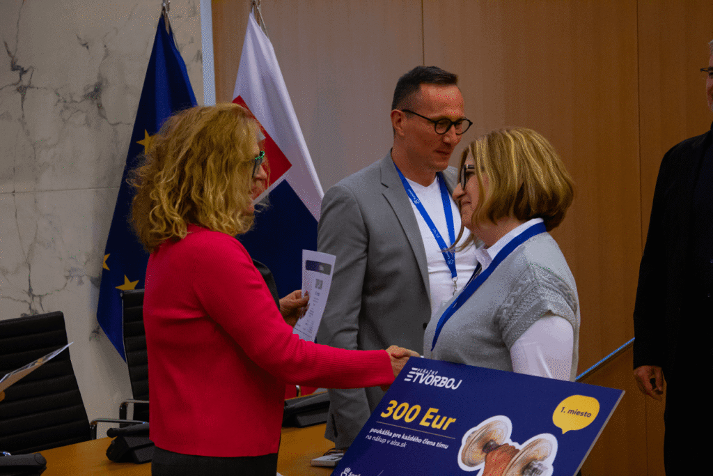 V miestnosti s vlajkami a mramorovou stenou stoja traja ľudia. Žena v ružovom podáva certifikát žene v okuliaroch a sivej veste, ktorá drží veľkú poukážku na 300 eur. Vedľa nich stojí muž v sivej bunde.