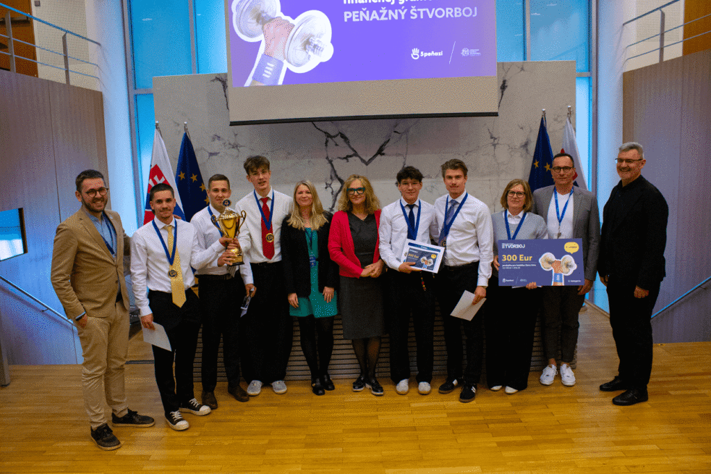 Skupina ľudí pózuje v interiéri na drevenej podlahe, niektorí držia trofej a veľký šek na 300 eur. Väčšina má na sebe medaily, stoja pred vlajkami a prezentačnou obrazovkou so slovenským textom.