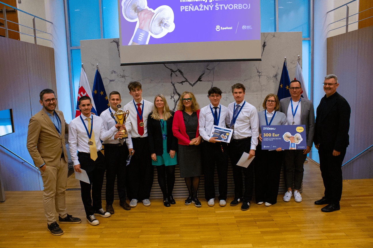 Skupina ľudí pózuje v interiéri na drevenej podlahe, niektorí držia trofej a veľký šek na 300 eur. Väčšina má na sebe medaily, stoja pred vlajkami a prezentačnou obrazovkou so slovenským textom.